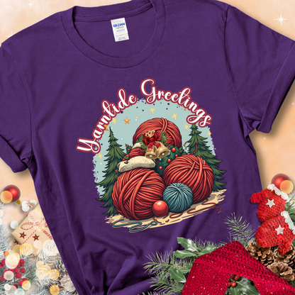 Purple t-shirt with red yarn balls, holiday greenery, bells, and the phrase “Yarntide Greetings.”