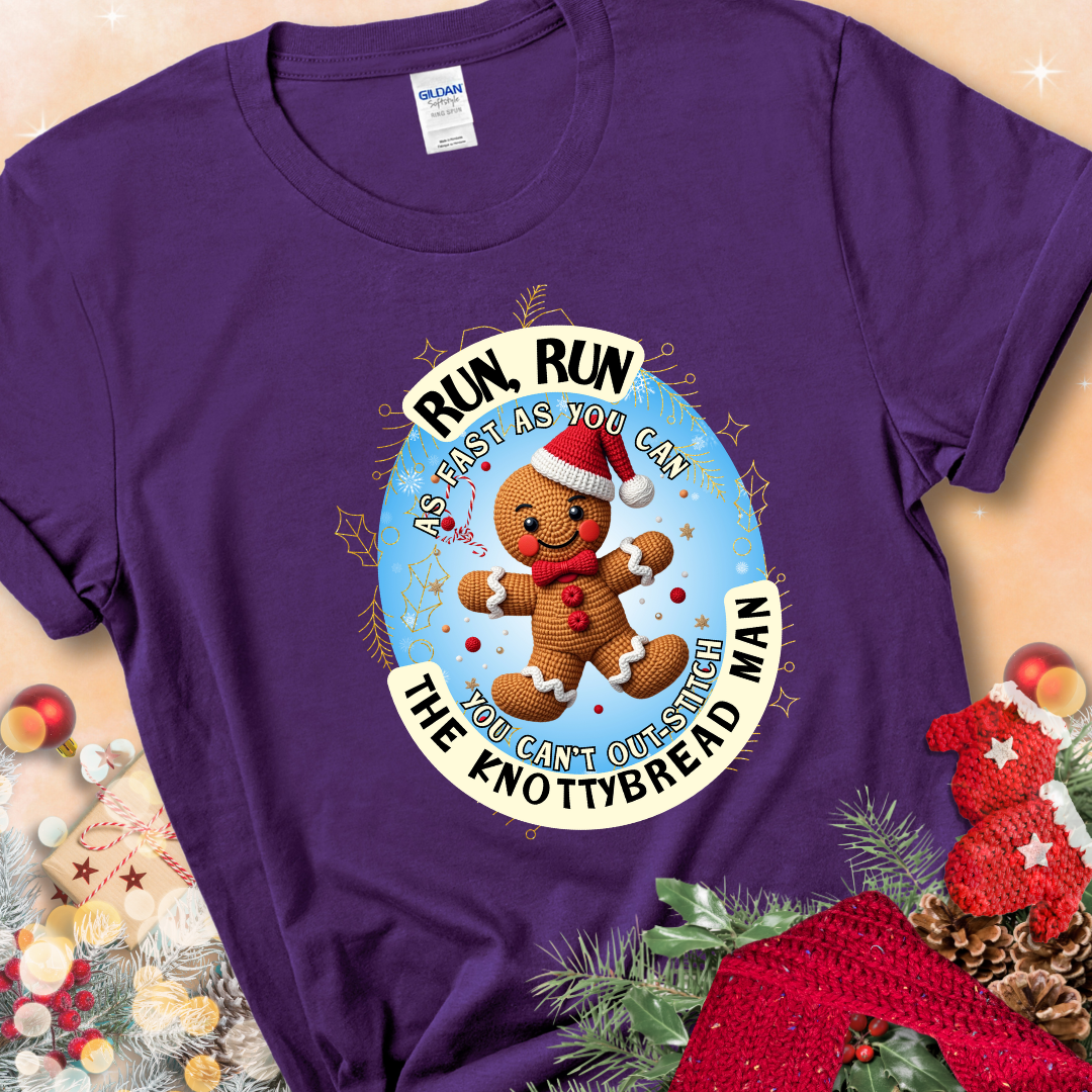 Purple t-shirt with a crocheted gingerbread man in a Santa hat and the phrase “You Can’t Out-Stitch the Knottybread Man.”