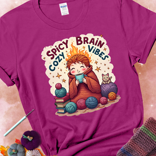 Antique Heliconia t-shirt with a red-haired person wrapped in a chunky crochet blanket, holding a mug, surrounded by yarn balls, books, a cat, and the phrase “Spicy Brain, Cozy Vibes.”