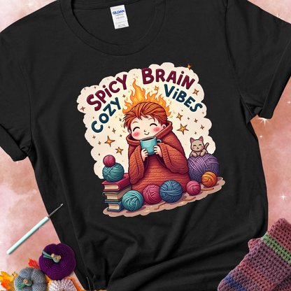 Black t-shirt with a red-haired person wrapped in a chunky crochet blanket, holding a mug, surrounded by yarn balls, books, a cat, and the phrase “Spicy Brain, Cozy Vibes.”