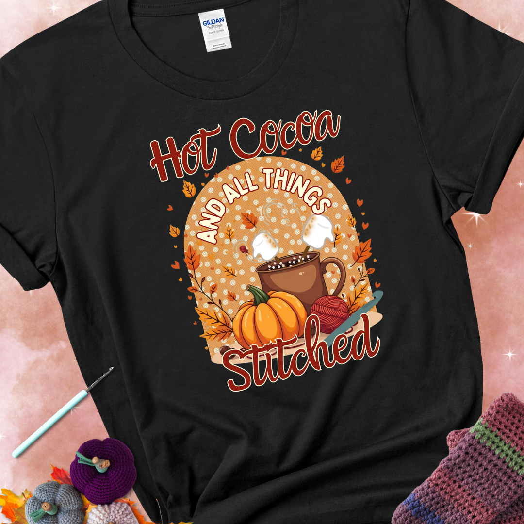 Black t-shirt with a cup of hot cocoa topped with marshmallows, a pumpkin, yarn balls, autumn leaves, and the phrase “Hot Cocoa & All Things Stitched.”