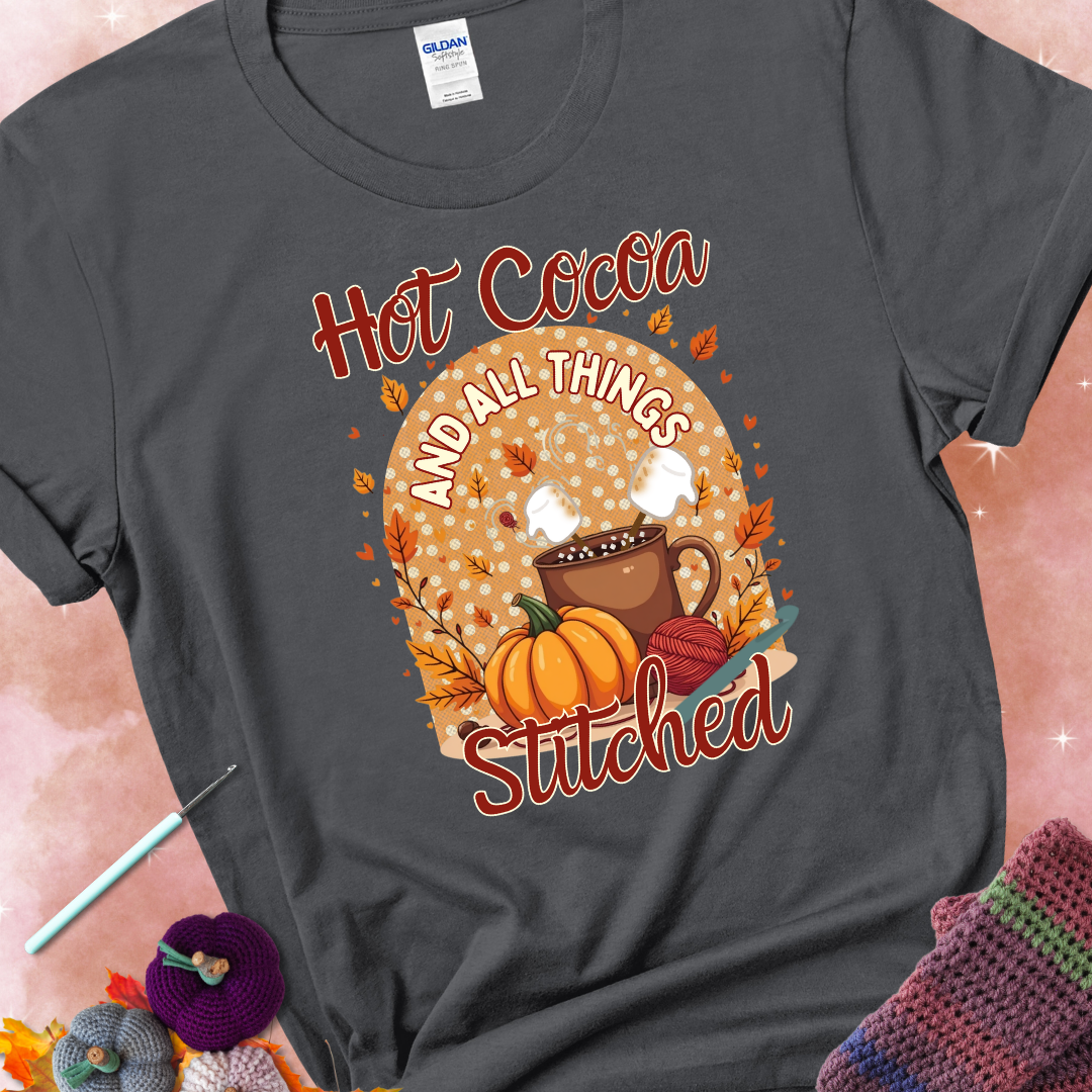 Charcoal t-shirt with a cup of hot cocoa topped with marshmallows, a pumpkin, yarn balls, autumn leaves, and the phrase “Hot Cocoa & All Things Stitched.”