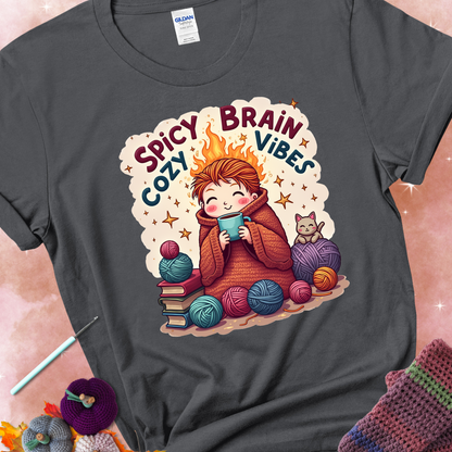 Charcoal t-shirt with a red-haired person wrapped in a chunky crochet blanket, holding a mug, surrounded by yarn balls, books, a cat, and the phrase “Spicy Brain, Cozy Vibes.”