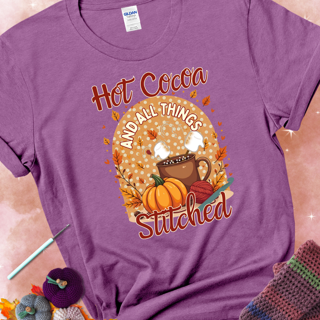 Heather Radiant Orchid t-shirt with a cup of hot cocoa topped with marshmallows, a pumpkin, yarn balls, autumn leaves, and the phrase “Hot Cocoa & All Things Stitched.”