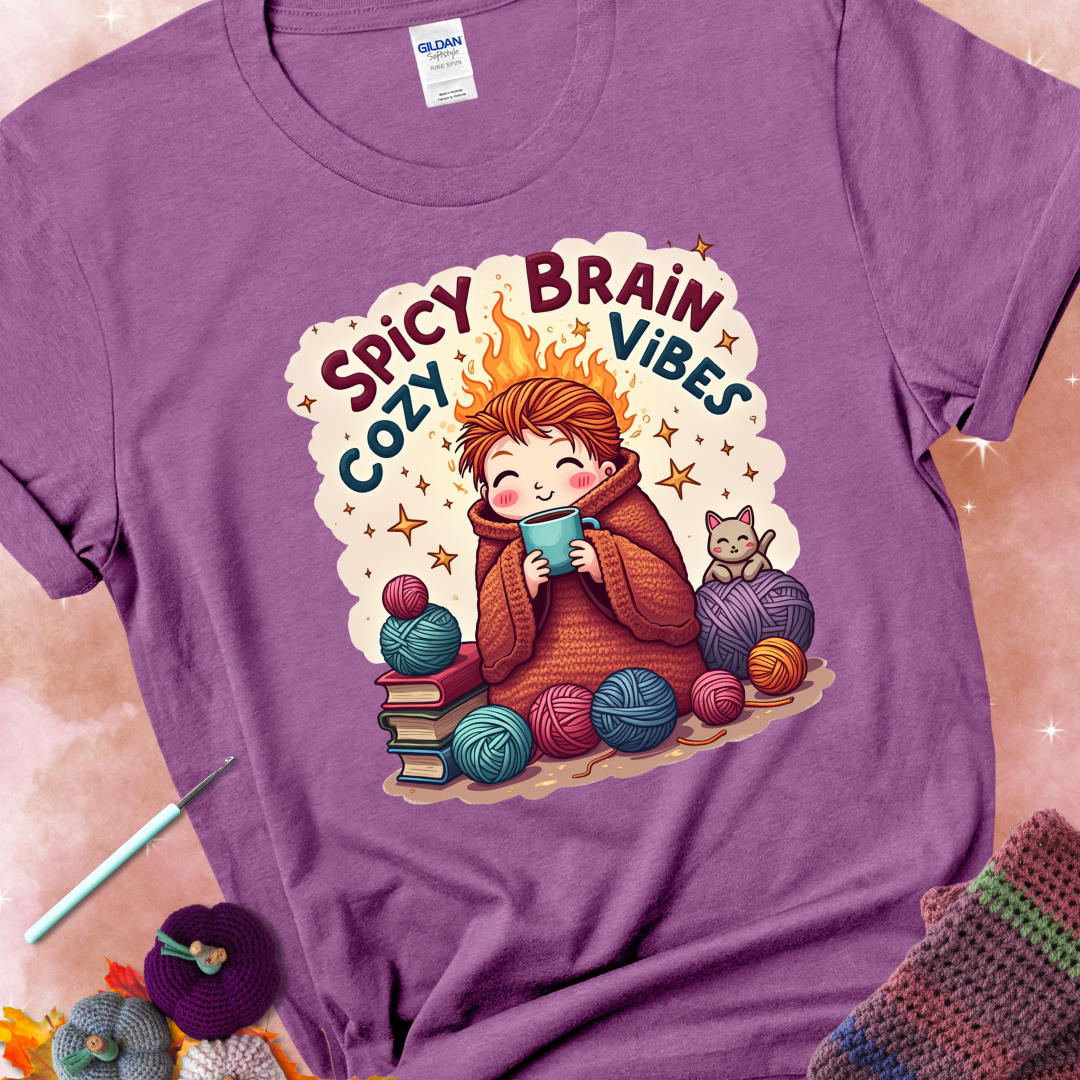 Heather Radiant Orchid t-shirt with a red-haired person wrapped in a chunky crochet blanket, holding a mug, surrounded by yarn balls, books, a cat, and the phrase “Spicy Brain, Cozy Vibes.”