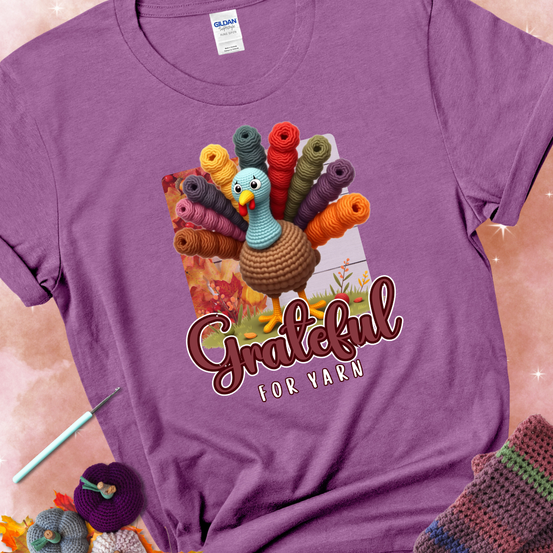 Heather radiant orchid t-shirt with a crocheted turkey, colorful yarn skeins for tail feathers, fall leaves, and the text “Grateful for Yarn.”