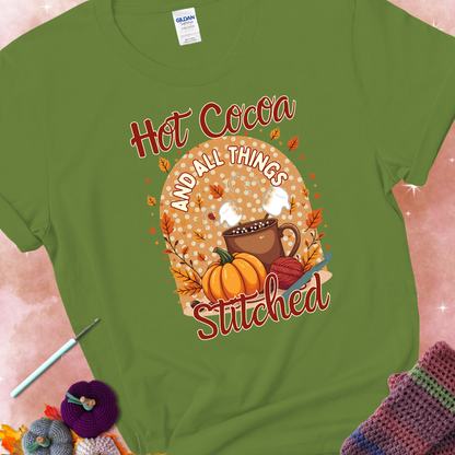 Kiwi t-shirt with a cup of hot cocoa topped with marshmallows, a pumpkin, yarn balls, autumn leaves, and the phrase “Hot Cocoa & All Things Stitched.”