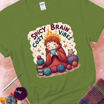 Kiwi t-shirt with a red-haired person wrapped in a chunky crochet blanket, holding a mug, surrounded by yarn balls, books, a cat, and the phrase “Spicy Brain, Cozy Vibes.”