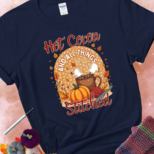 Navy t-shirt with a cup of hot cocoa topped with marshmallows, a pumpkin, yarn balls, autumn leaves, and the phrase “Hot Cocoa & All Things Stitched.”