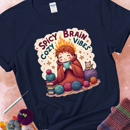 Navy t-shirt with a red-haired person wrapped in a chunky crochet blanket, holding a mug, surrounded by yarn balls, books, a cat, and the phrase “Spicy Brain, Cozy Vibes.”