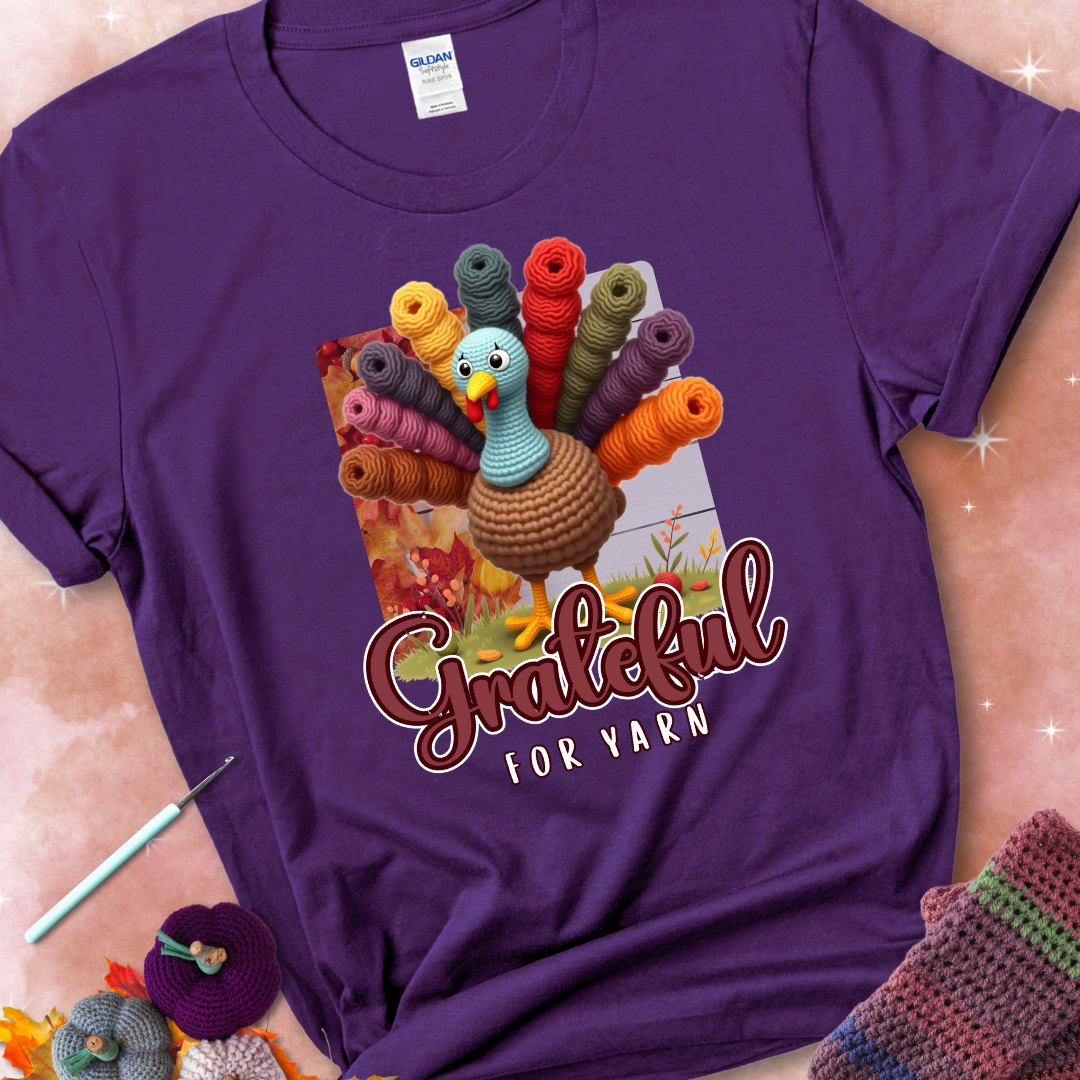 Purple t-shirt with a crocheted turkey, colorful yarn skeins for tail feathers, fall leaves, and the text “Grateful for Yarn.”