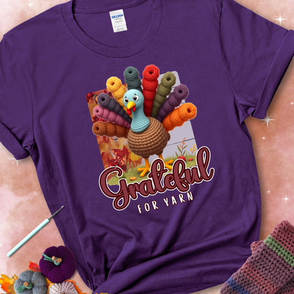 Purple t-shirt with a crocheted turkey, colorful yarn skeins for tail feathers, fall leaves, and the text “Grateful for Yarn.”