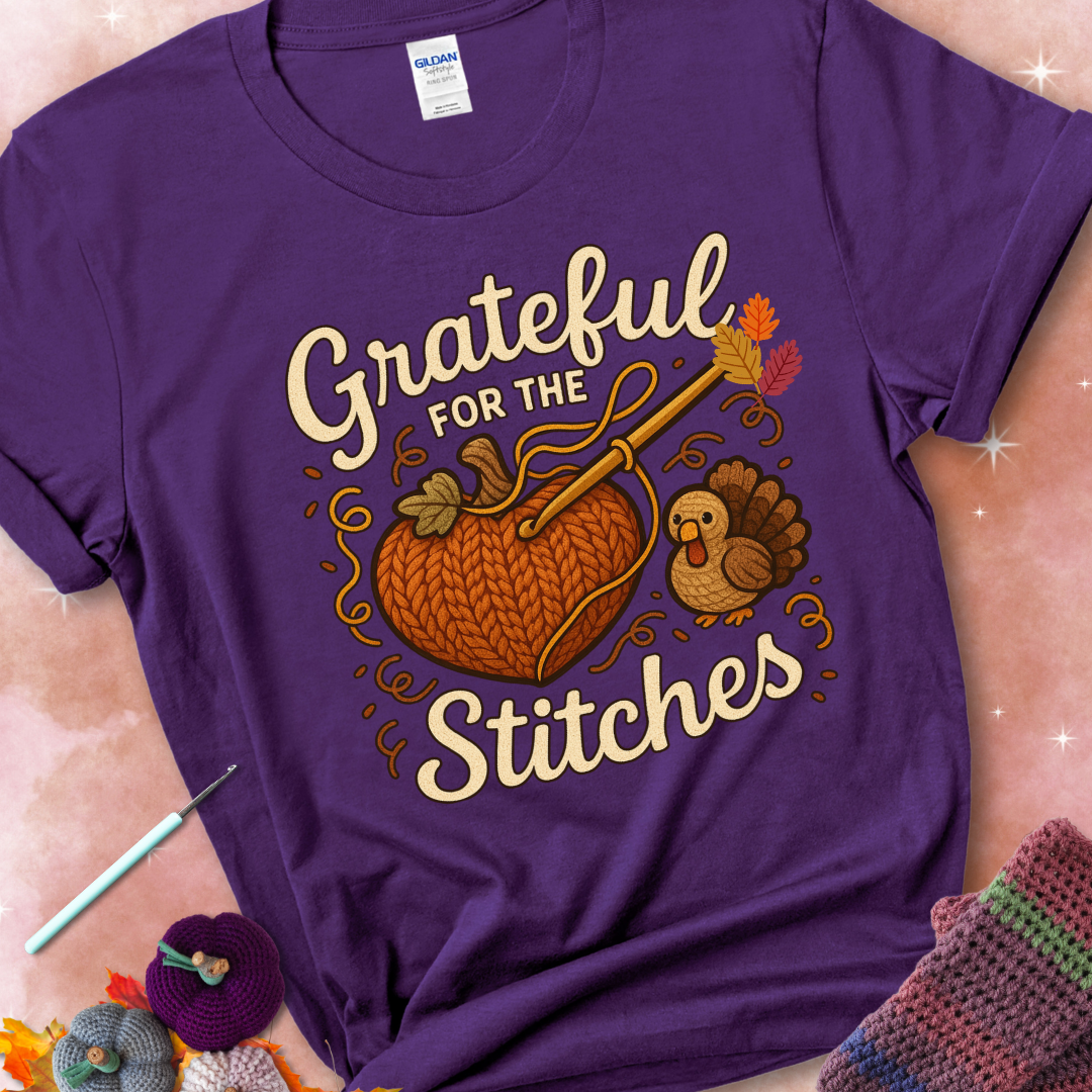 Purple t-shirt with a crochet hook stitching a heart-shaped pumpkin, surrounded by yarn and a small turkey, with the phrase “Grateful for the Stitches.”