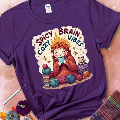 Purple t-shirt with a red-haired person wrapped in a chunky crochet blanket, holding a mug, surrounded by yarn balls, books, a cat, and the phrase “Spicy Brain, Cozy Vibes.”