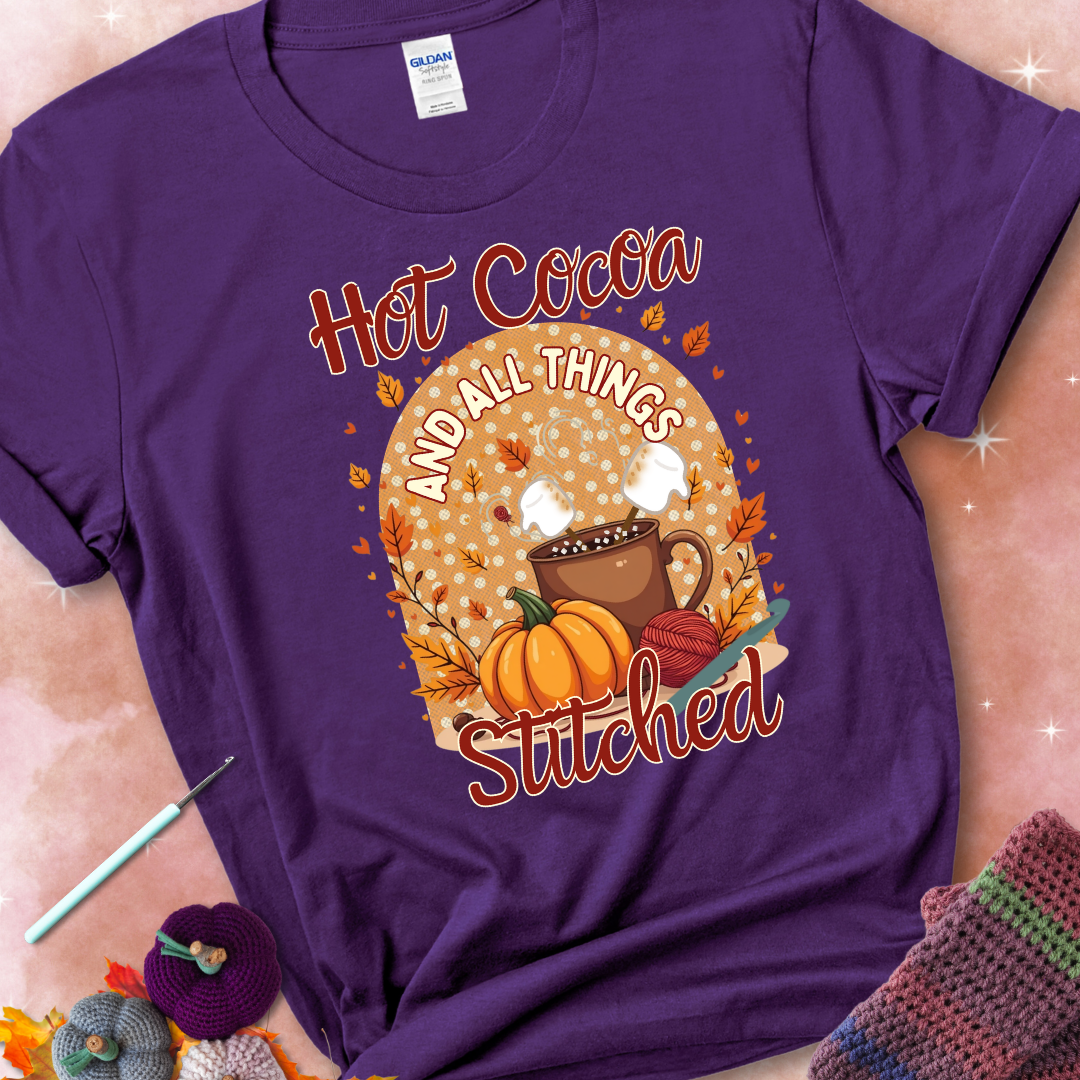 Purple t-shirt with a cup of hot cocoa topped with marshmallows, a pumpkin, yarn balls, autumn leaves, and the phrase “Hot Cocoa & All Things Stitched.”
