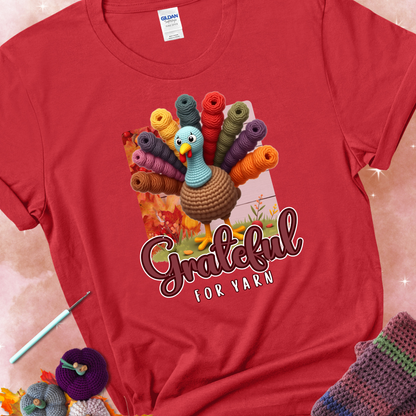 Red t-shirt with a crocheted turkey, colorful yarn skeins for tail feathers, fall leaves, and the text “Grateful for Yarn.”