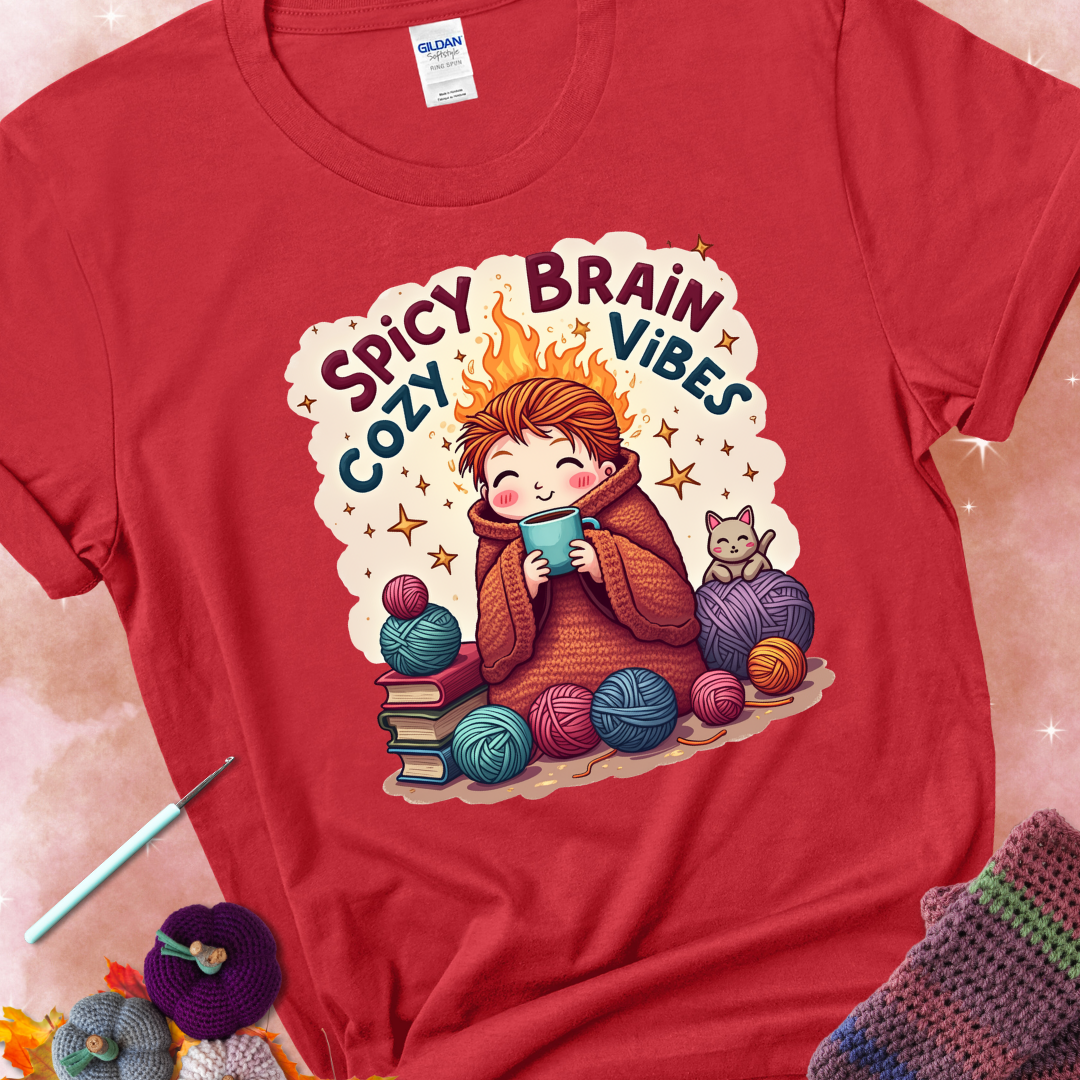 Red t-shirt with a red-haired person wrapped in a chunky crochet blanket, holding a mug, surrounded by yarn balls, books, a cat, and the phrase “Spicy Brain, Cozy Vibes.”