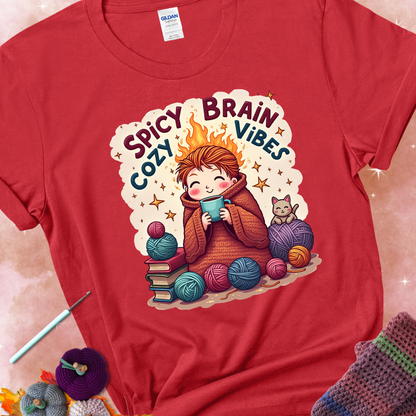 Red t-shirt with a red-haired person wrapped in a chunky crochet blanket, holding a mug, surrounded by yarn balls, books, a cat, and the phrase “Spicy Brain, Cozy Vibes.”