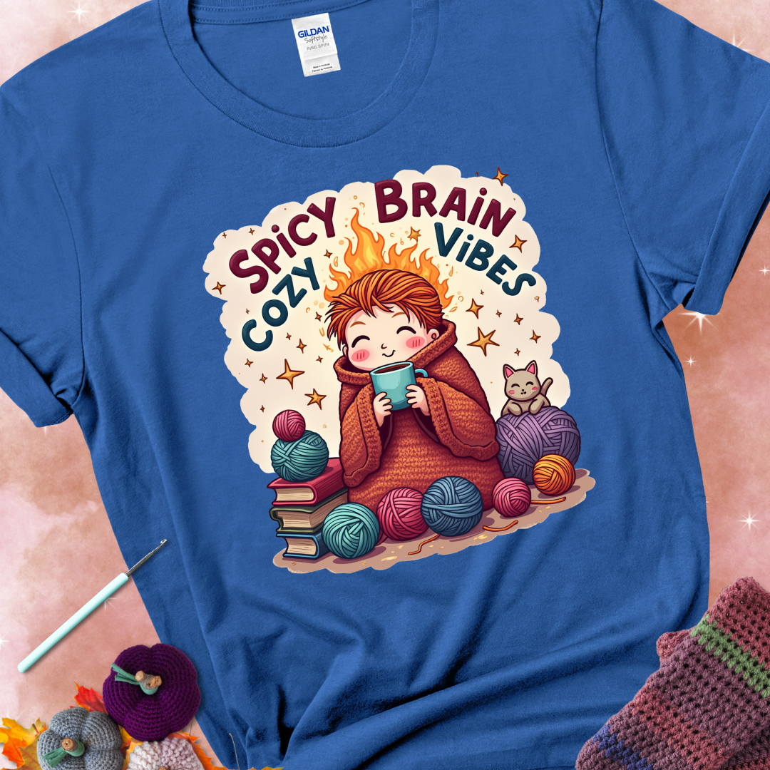 Royal t-shirt with a red-haired person wrapped in a chunky crochet blanket, holding a mug, surrounded by yarn balls, books, a cat, and the phrase “Spicy Brain, Cozy Vibes.”