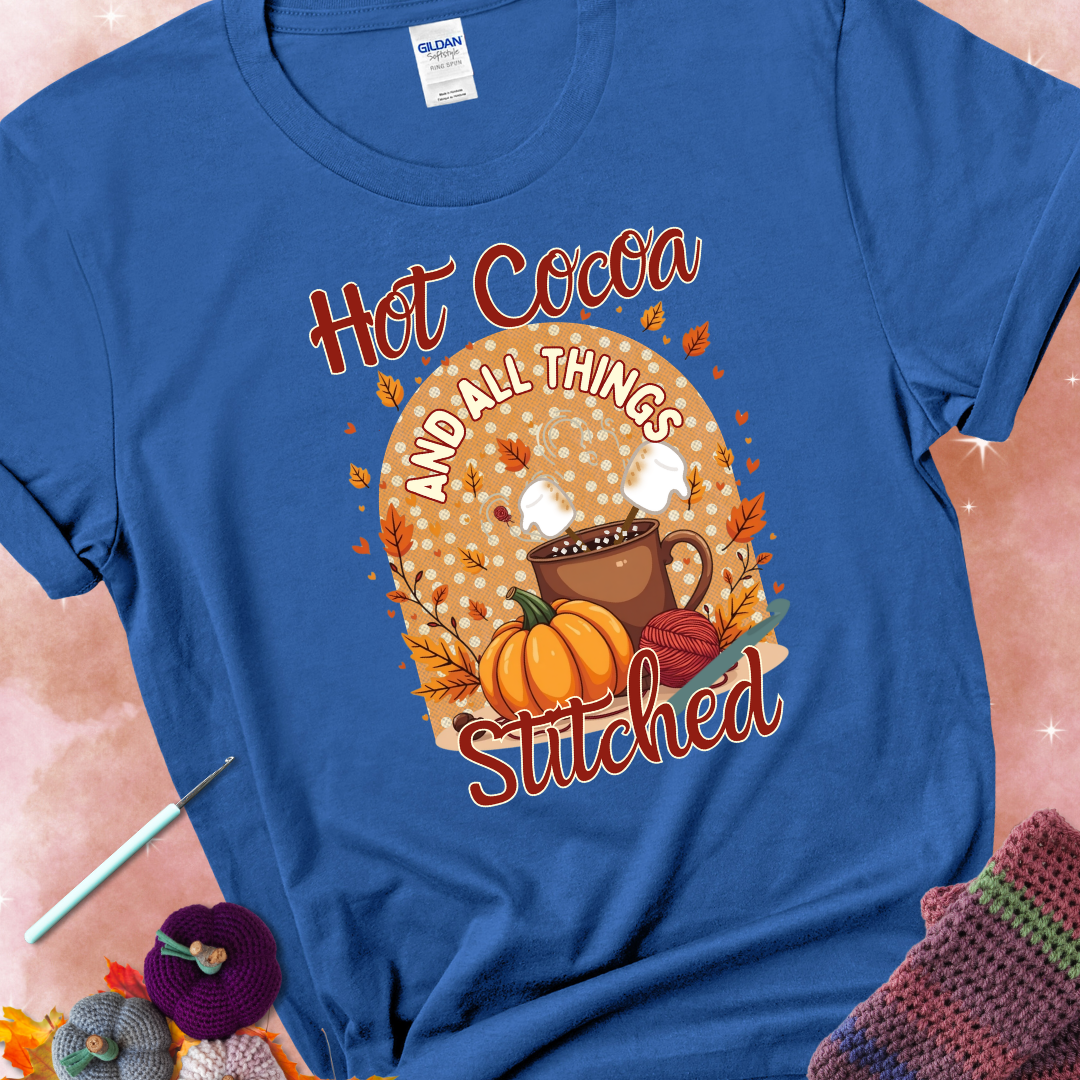 Royal t-shirt with a cup of hot cocoa topped with marshmallows, a pumpkin, yarn balls, autumn leaves, and the phrase “Hot Cocoa & All Things Stitched.”