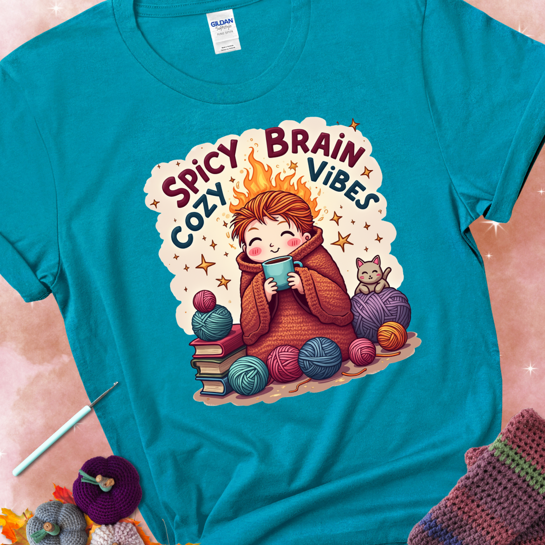 Tropical Blue t-shirt with a red-haired person wrapped in a chunky crochet blanket, holding a mug, surrounded by yarn balls, books, a cat, and the phrase “Spicy Brain, Cozy Vibes.”
