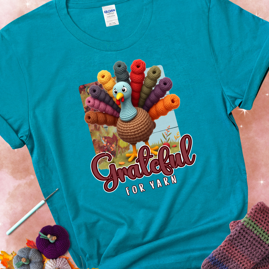 Tropical blue t-shirt with a crocheted turkey, colorful yarn skeins for tail feathers, fall leaves, and the text “Grateful for Yarn.”