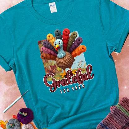 Tropical blue t-shirt with a crocheted turkey, colorful yarn skeins for tail feathers, fall leaves, and the text “Grateful for Yarn.”