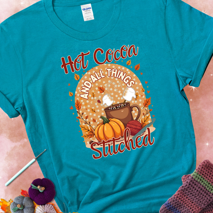 Tropical Blue t-shirt with a cup of hot cocoa topped with marshmallows, a pumpkin, yarn balls, autumn leaves, and the phrase “Hot Cocoa & All Things Stitched.”