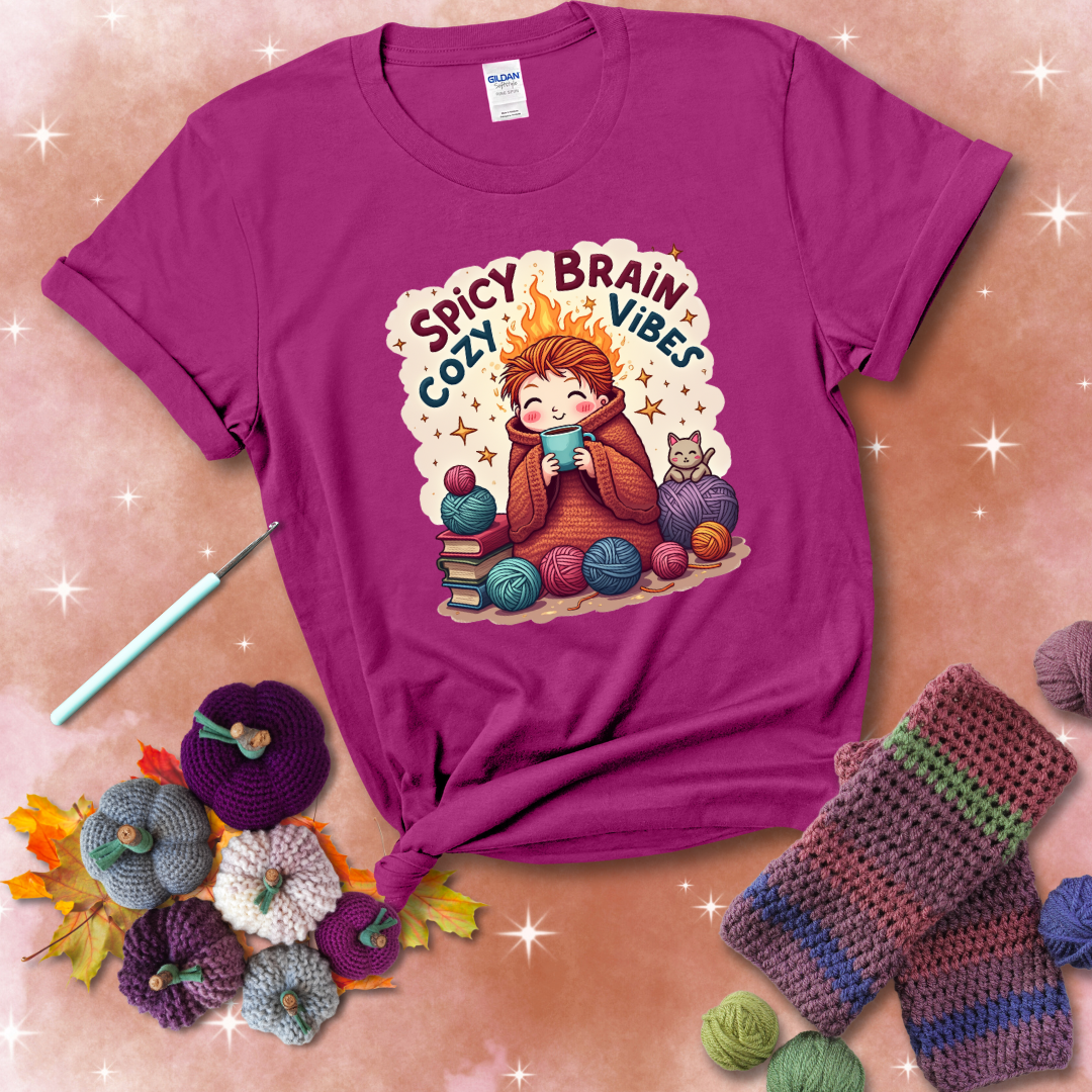 A person with flaming red hair wrapped in a chunky crochet blanket, holding a mug, surrounded by yarn balls, books, a cat, and the phrase "Spicy Brain Cozy Vibes" showing a magenta tee with a festive Fall background