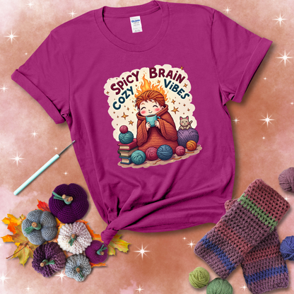 A person with flaming red hair wrapped in a chunky crochet blanket, holding a mug, surrounded by yarn balls, books, a cat, and the phrase "Spicy Brain Cozy Vibes" showing a magenta tee with a festive Fall background