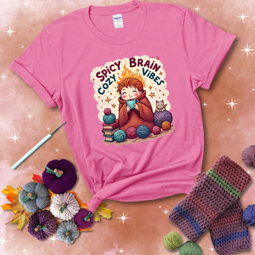 A person with flaming red hair wrapped in a chunky crochet blanket, holding a mug, surrounded by yarn balls, books, a cat, and the phrase "Spicy Brain Cozy Vibes" showing a pink tee with a festive Fall background