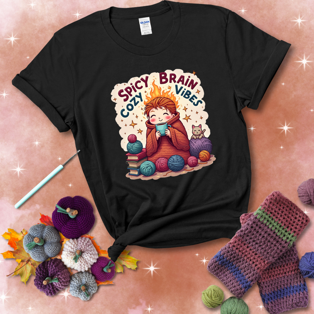 A person with flaming red hair wrapped in a chunky crochet blanket, holding a mug, surrounded by yarn balls, books, a cat, and the phrase "Spicy Brain Cozy Vibes" showing a black tee with a festive Fall background