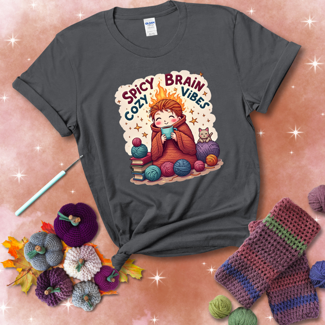 A person with flaming red hair wrapped in a chunky crochet blanket, holding a mug, surrounded by yarn balls, books, a cat, and the phrase "Spicy Brain Cozy Vibes" showing a dark grey tee with a festive Fall background