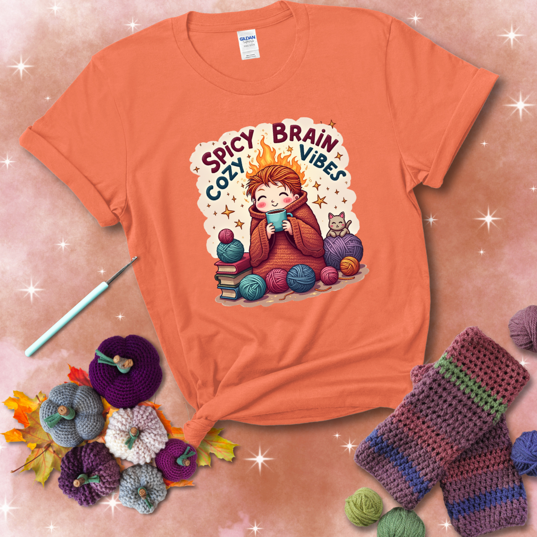 A person with flaming red hair wrapped in a chunky crochet blanket, holding a mug, surrounded by yarn balls, books, a cat, and the phrase "Spicy Brain Cozy Vibes" showing a light orange tee with a festive Fall background