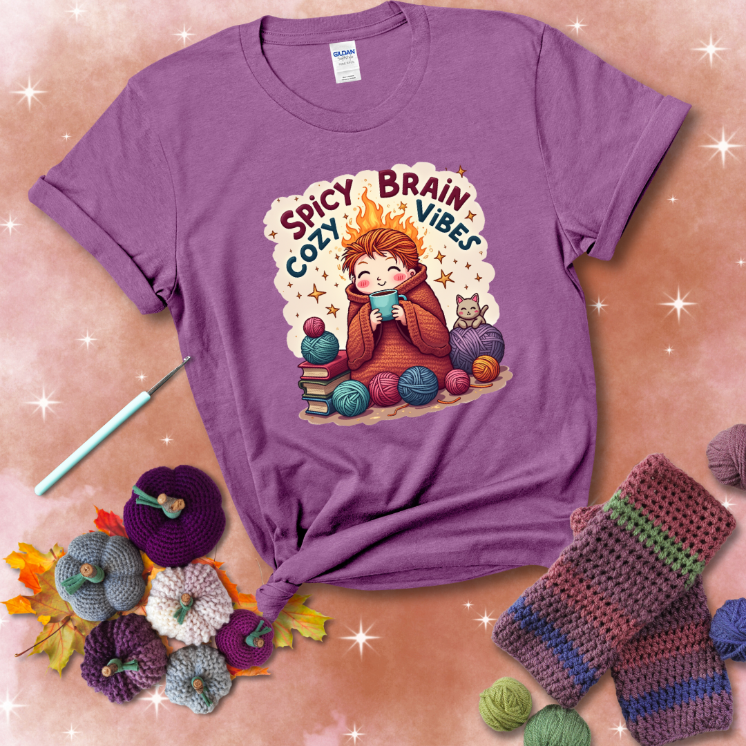A person with flaming red hair wrapped in a chunky crochet blanket, holding a mug, surrounded by yarn balls, books, a cat, and the phrase "Spicy Brain Cozy Vibes" showing a light purple tee with a festive Fall background