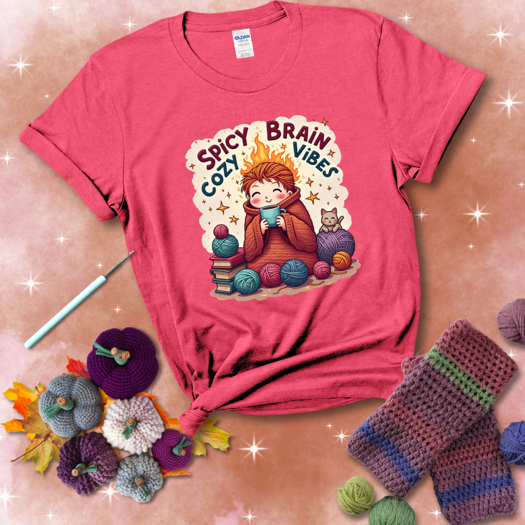 A person with flaming red hair wrapped in a chunky crochet blanket, holding a mug, surrounded by yarn balls, books, a cat, and the phrase "Spicy Brain Cozy Vibes" showing a light red tee with a festive Fall background
