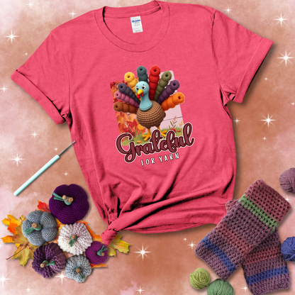 A crocheted turkey with colorful yarn skeins for tail feathers, standing on fall leaves with the text “Grateful for Yarn” showing a light red tee with a festive Fall background