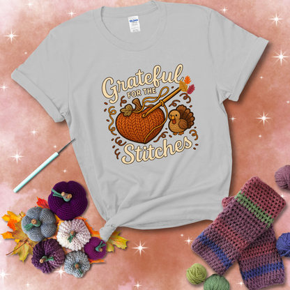 A cozy fall scene with a crochet hook stitching a heart-shaped pumpkin, surrounded by yarn and a small turkey, showing a light grey tee with a festive Fall background
