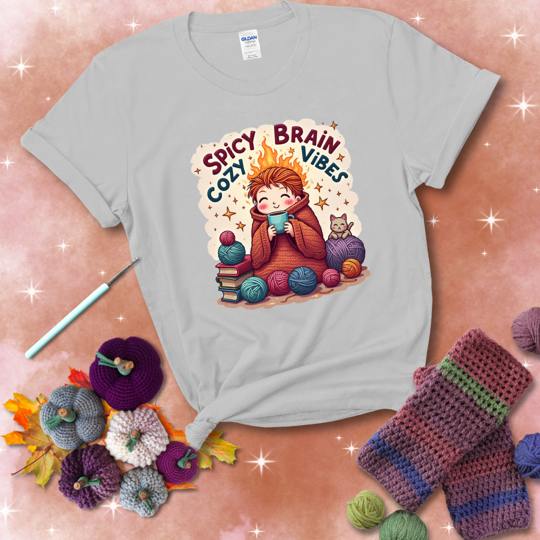 A person with flaming red hair wrapped in a chunky crochet blanket, holding a mug, surrounded by yarn balls, books, a cat, and the phrase "Spicy Brain Cozy Vibes" showing a light grey tee with a festive Fall background
