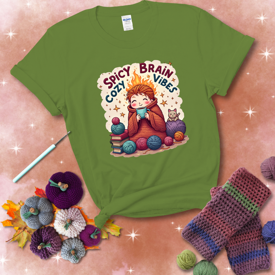 A person with flaming red hair wrapped in a chunky crochet blanket, holding a mug, surrounded by yarn balls, books, a cat, and the phrase "Spicy Brain Cozy Vibes" showing a bright green tee with a festive Fall background