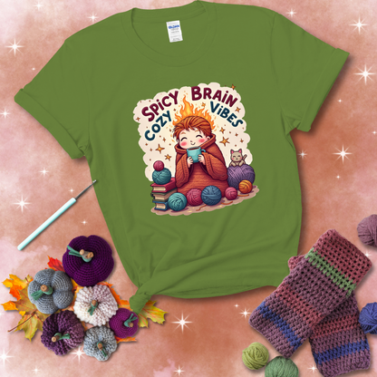 A person with flaming red hair wrapped in a chunky crochet blanket, holding a mug, surrounded by yarn balls, books, a cat, and the phrase "Spicy Brain Cozy Vibes" showing a bright green tee with a festive Fall background
