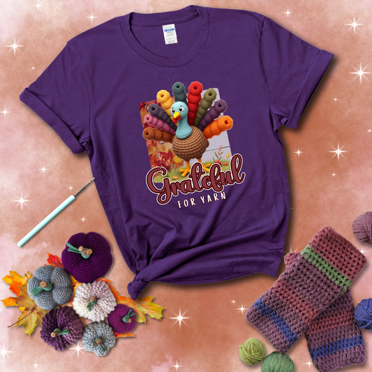 A crocheted turkey with colorful yarn skeins for tail feathers, standing on fall leaves with the text “Grateful for Yarn” showing a purple tee with a festive Fall background