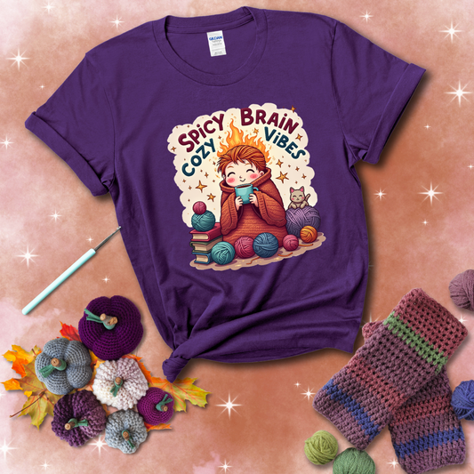 A person with flaming red hair wrapped in a chunky crochet blanket, holding a mug, surrounded by yarn balls, books, a cat, and the phrase "Spicy Brain Cozy Vibes" showing a purple tee with a festive Fall background