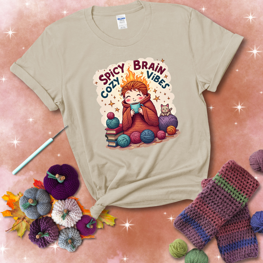 A person with flaming red hair wrapped in a chunky crochet blanket, holding a mug, surrounded by yarn balls, books, a cat, and the phrase "Spicy Brain Cozy Vibes" showing a beige tee with a festive Fall background