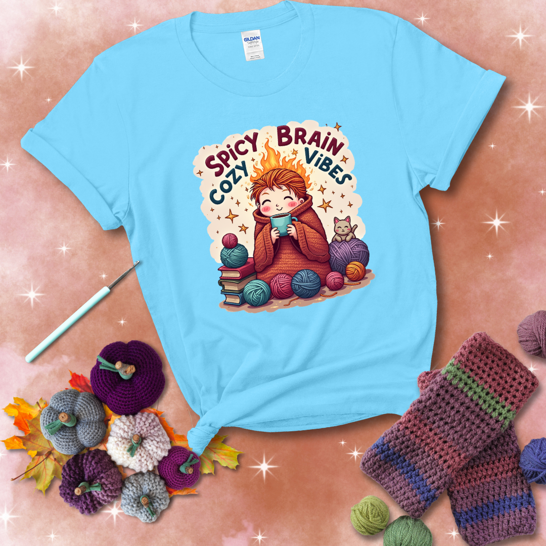 A person with flaming red hair wrapped in a chunky crochet blanket, holding a mug, surrounded by yarn balls, books, a cat, and the phrase "Spicy Brain Cozy Vibes" showing a light blue tee with a festive Fall background