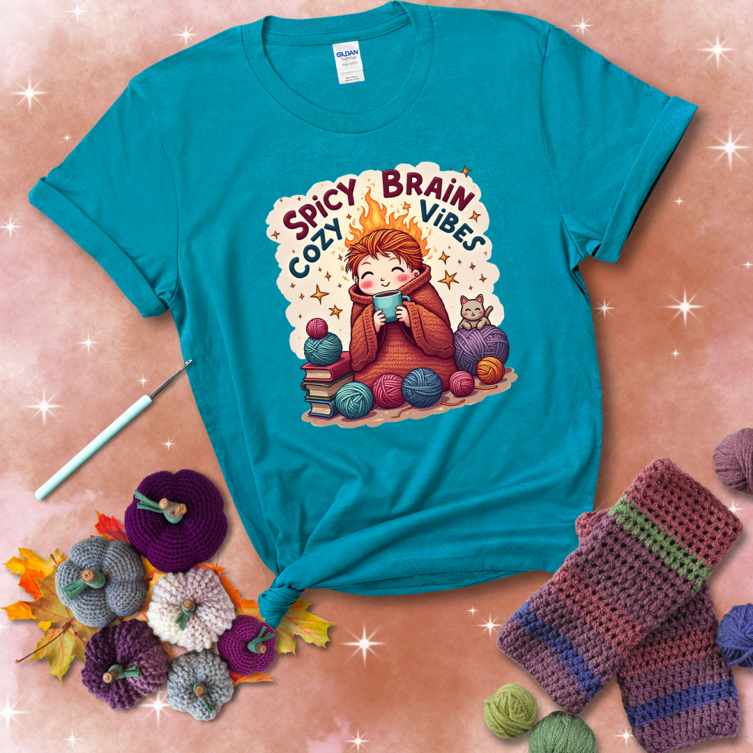A person with flaming red hair wrapped in a chunky crochet blanket, holding a mug, surrounded by yarn balls, books, a cat, and the phrase "Spicy Brain Cozy Vibes" showing a blue-green tee with a festive Fall background