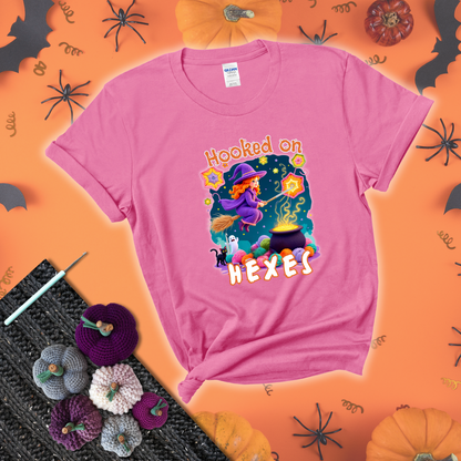 Witchy vibe with stars, hexagons, and a crochet hook. Text reads: “Hooked on Hexes.” A mystical and moody tee for fans of spooky stitches showing a pink tee with a festive Halloween background