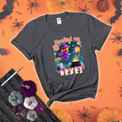 Witchy vibe with stars, hexagons, and a crochet hook. Text reads: “Hooked on Hexes.” A mystical and moody tee for fans of spooky stitches showing a dark grey tee with a festive Halloween background