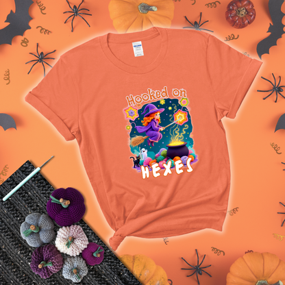 Witchy vibe with stars, hexagons, and a crochet hook. Text reads: “Hooked on Hexes.” A mystical and moody tee for fans of spooky stitches showing a light orange tee with a festive Halloween background