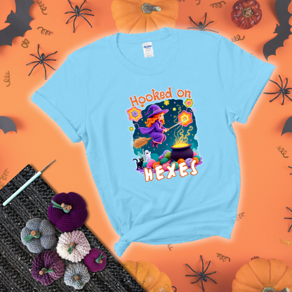 Witchy vibe with stars, hexagons, and a crochet hook. Text reads: “Hooked on Hexes.” A mystical and moody tee for fans of spooky stitches showing a light blue tee with a festive Halloween background